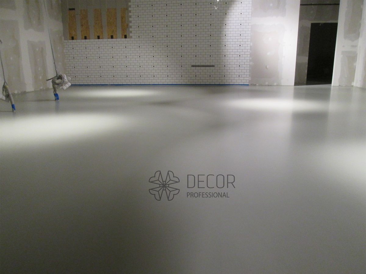 Jasnoszara, gładka posadzka w pomieszczeniu z białą cegłą na ścianie. Widoczne narzędzia malarskie i logo firmy Decor Professional.