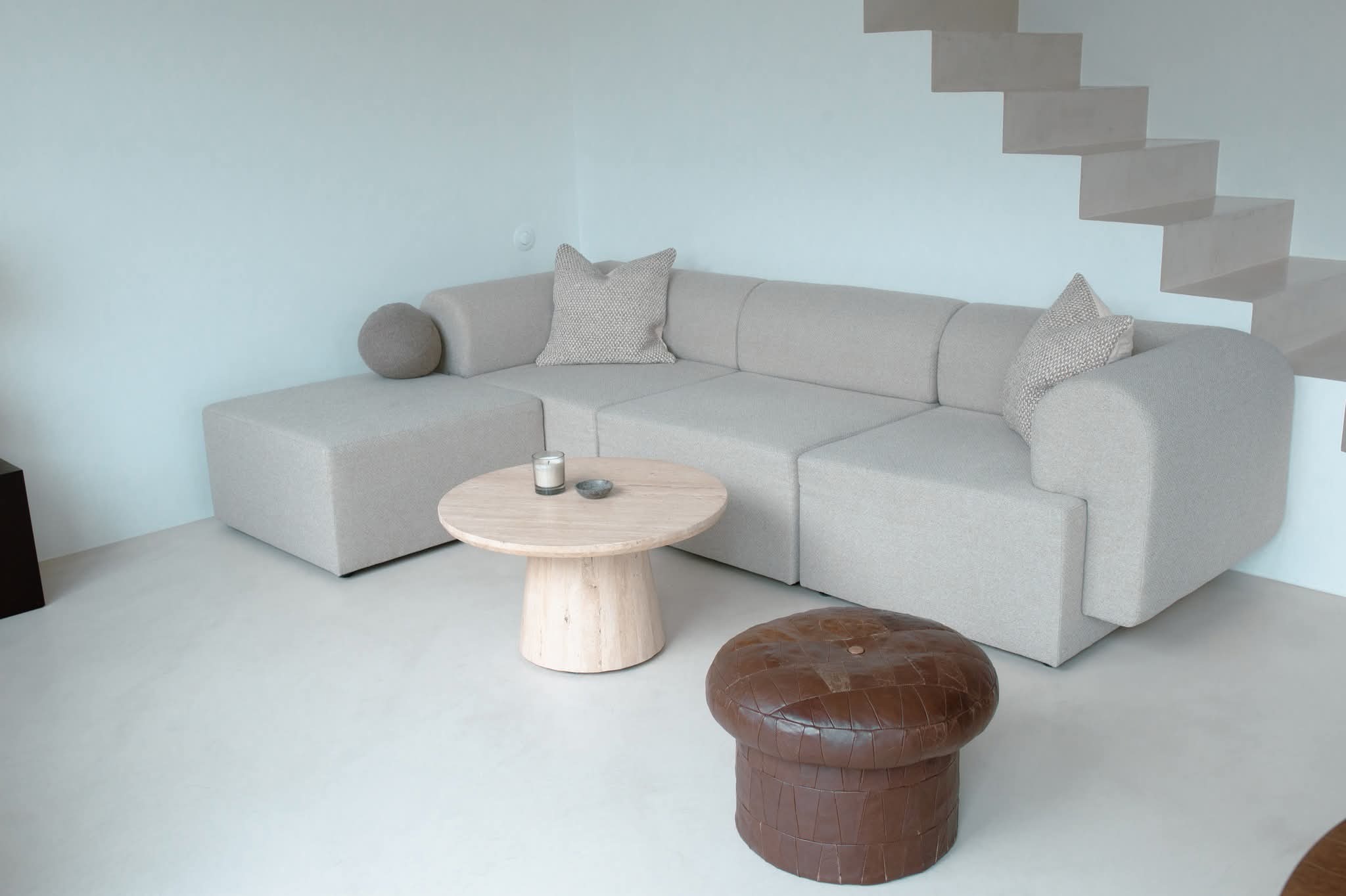 Jasnoszara, modułowa sofa narożna z poduszkami, okrągły stolik kawowy i pufa na jasnej posadzce. W tle minimalistyczne schody i ściana w odcieniach bieli.