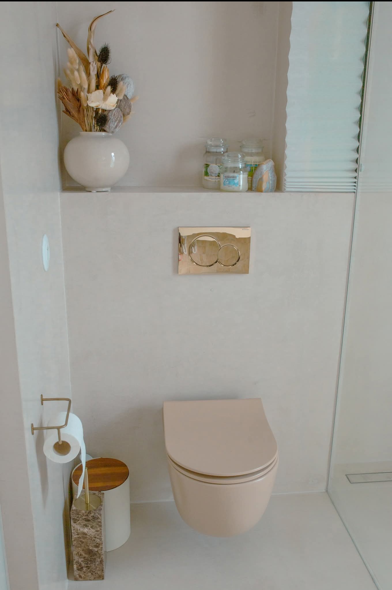 Beżowa toaleta podtynkowa z złotym przyciskiem spłukującym, w łazience wykończonej mikrocementem. Na półce dekoracje: suszone kwiaty i świece.