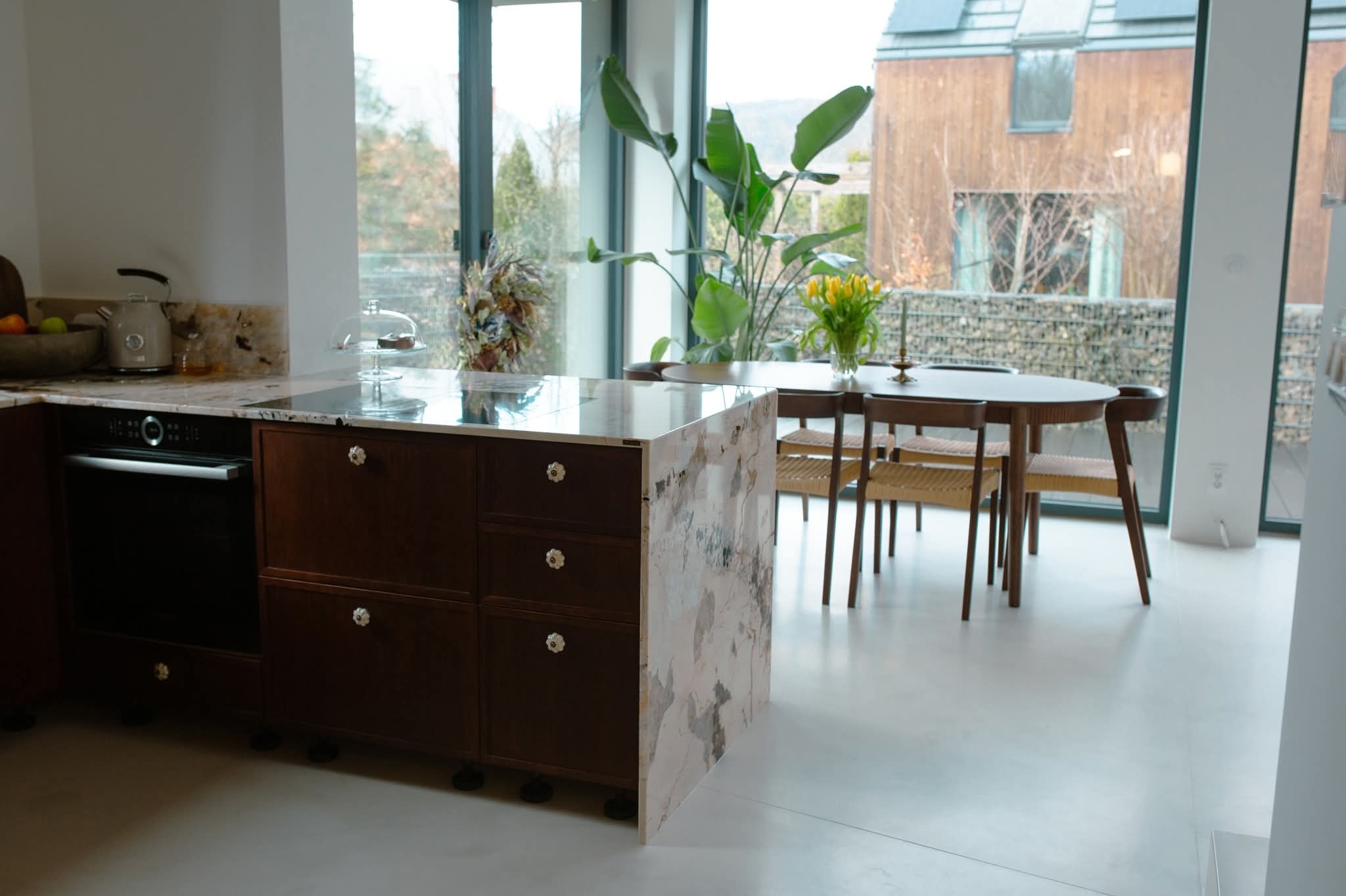 Jasna, gładka posadzka w nowoczesnej kuchni z ciemnymi meblami i marmurowym blatem. Duże okno z widokiem na ogród. Minimalistyczny design.