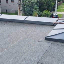 Łukasz pasamonik - Szary dach pokryty papą z obróbkami blacharskimi w kolorze grafitowym. W tle widoczna ulica i fragment budynku.