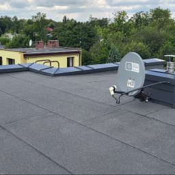 Łukasz pasamonik - Szary dach pokryty papą termozgrzewalną z widocznymi obróbkami blacharskimi, kominami i anteną satelitarną. W tle zabudowa miejska i zieleń.