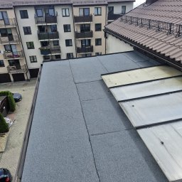 Łukasz pasamonik - Szary, płaski dach pokryty papą termozgrzewalną, z widocznymi łączeniami, sąsiadujący z dachem skośnym z brązowej blachodachówki i przezroczystymi panelami. W tle budynki mieszkalne.