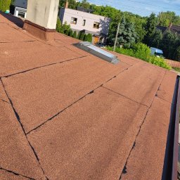 Łukasz pasamonik - Dach pokryty papą termozgrzewalną w odcieniach brązu, z widocznym kominem i oknem dachowym. W tle zabudowa jednorodzinna i błękitne niebo z chmurami.