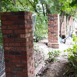 Łukasz pasamonik - Dwie murowane kolumny z czerwonej cegły na betonowej podstawie, otoczone zielenią drzew i krzewów. Widoczne ślady prac ziemnych.