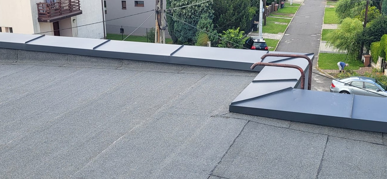 Szary dach pokryty papą z obróbkami blacharskimi w kolorze grafitowym. W tle widoczna ulica i fragment budynku.