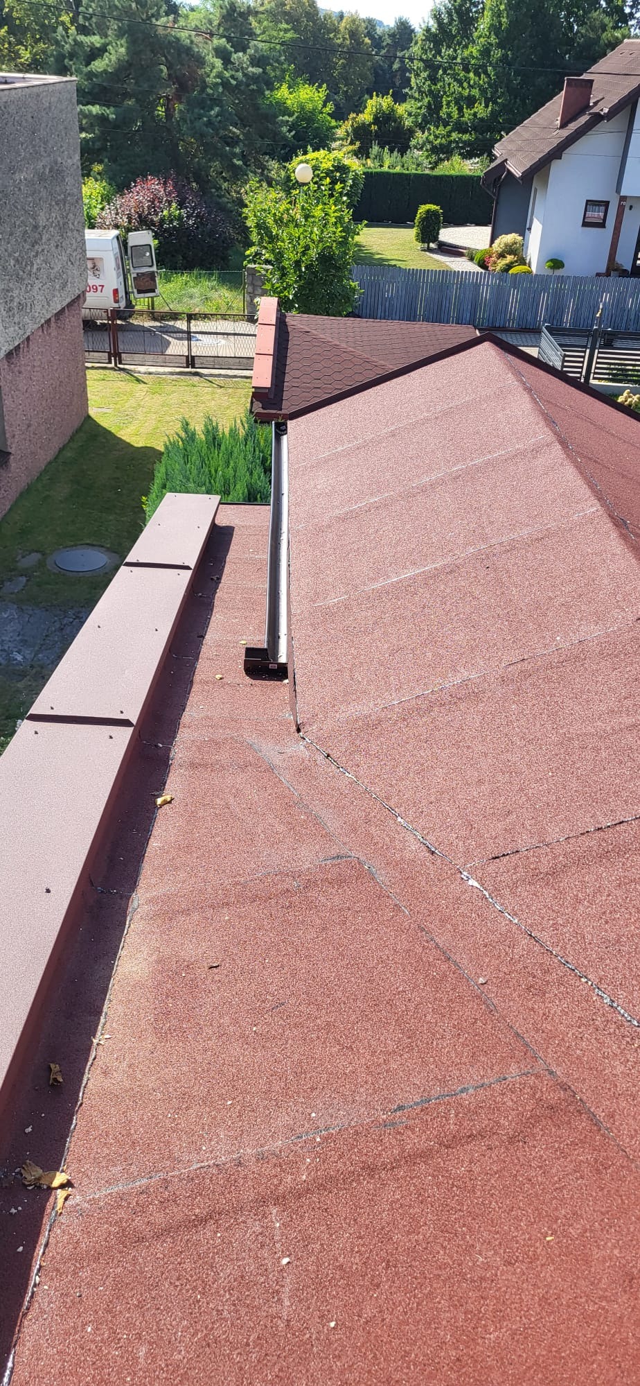 Czerwony dach z papy bitumicznej z widoczną rynną i obróbką blacharską, na tle zieleni i zabudowań. Widok z góry.