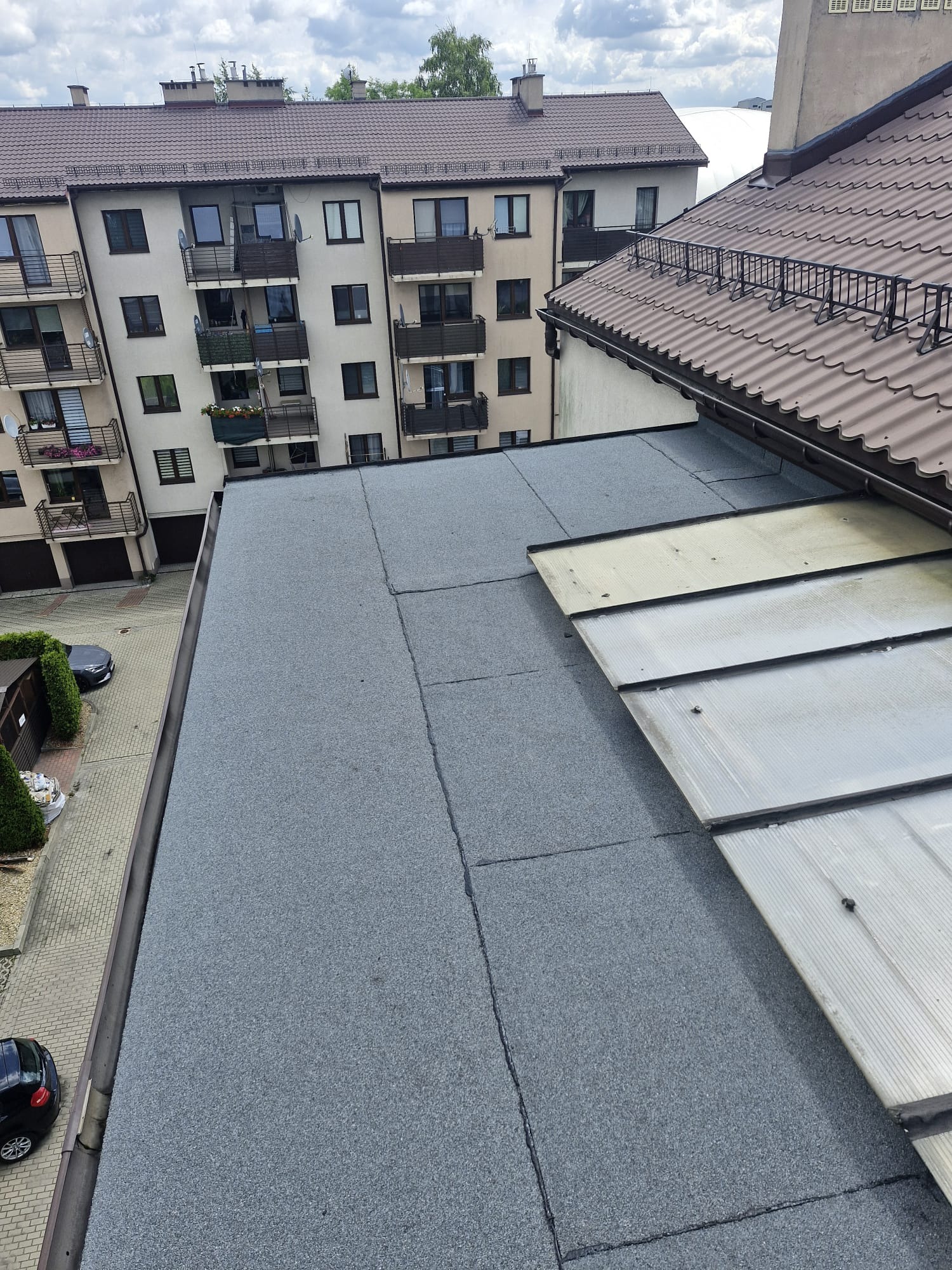 Szary, płaski dach pokryty papą termozgrzewalną, z widocznymi łączeniami, sąsiadujący z dachem skośnym z brązowej blachodachówki i przezroczystymi panelami. W tle budynki mieszkalne.