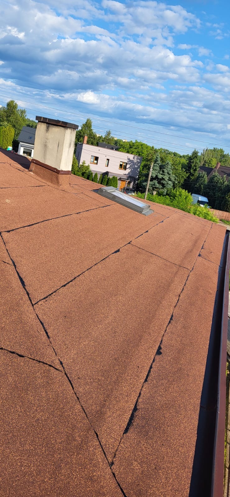 Dach pokryty papą termozgrzewalną w odcieniach brązu, z widocznym kominem i oknem dachowym. W tle zabudowa jednorodzinna i błękitne niebo z chmurami.