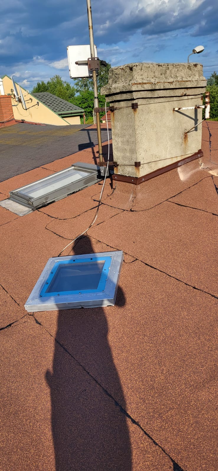 Dach pokryty papą z dwoma oknami dachowymi i kominem. Widoczny cień osoby. Jeden z okien jest otwarty. Na dachu widoczna instalacja odgromowa.