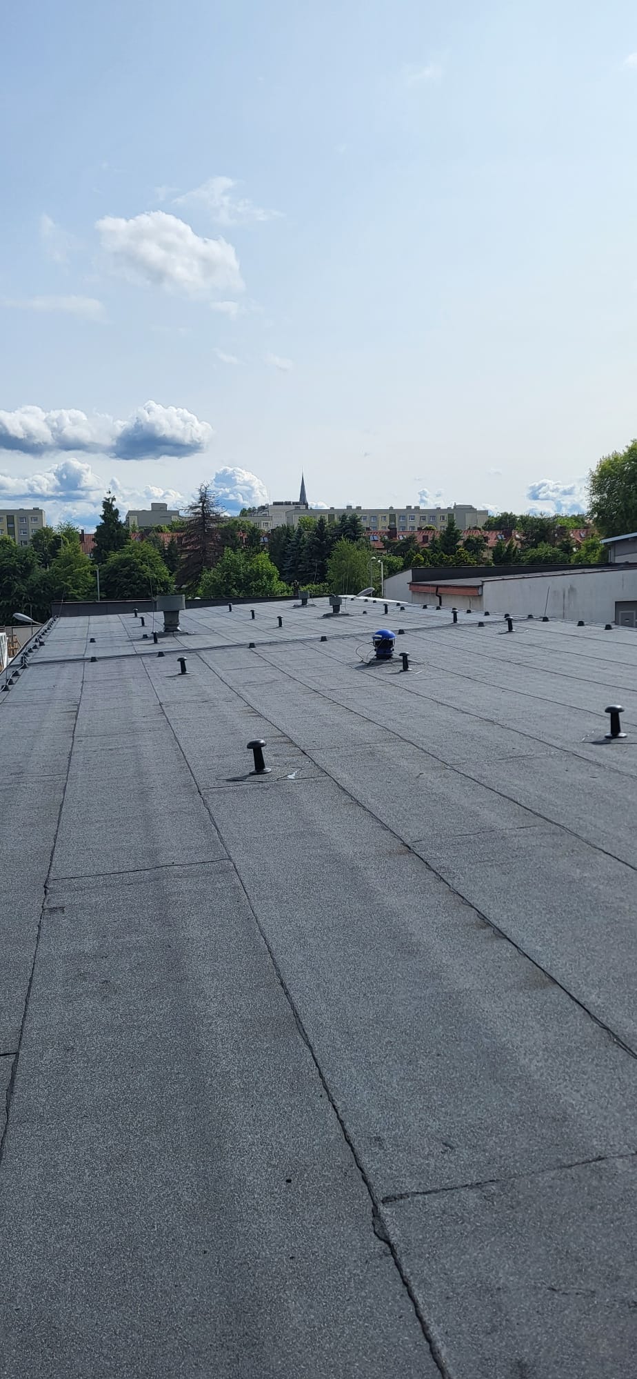 Płaski dach pokryty papą termozgrzewalną z kominkami wentylacyjnymi na tle zabudowy miejskiej i błękitnego nieba z chmurami.