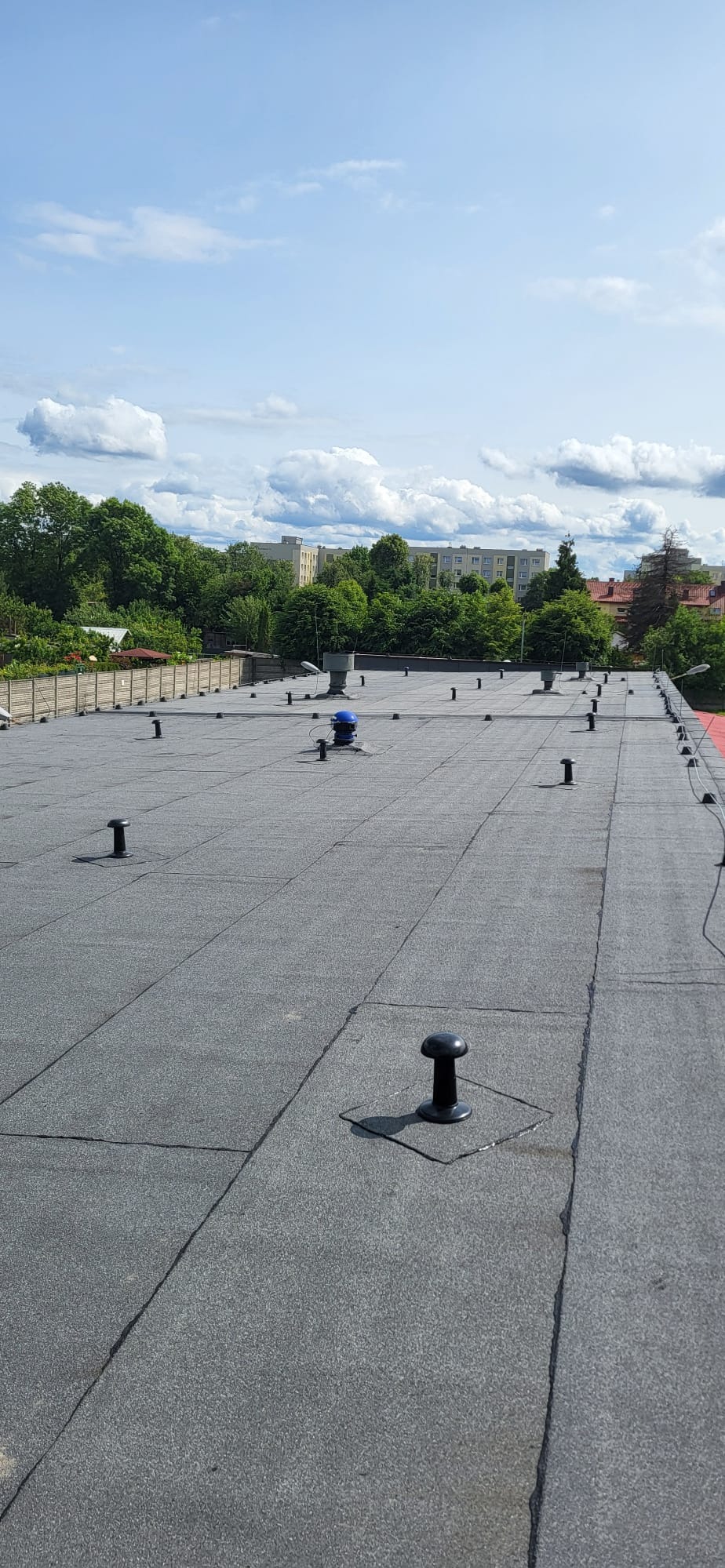 Szary dach pokryty papą termozgrzewalną z kominkami wentylacyjnymi na tle błękitnego nieba z chmurami i zieleni drzew. Widok z góry.