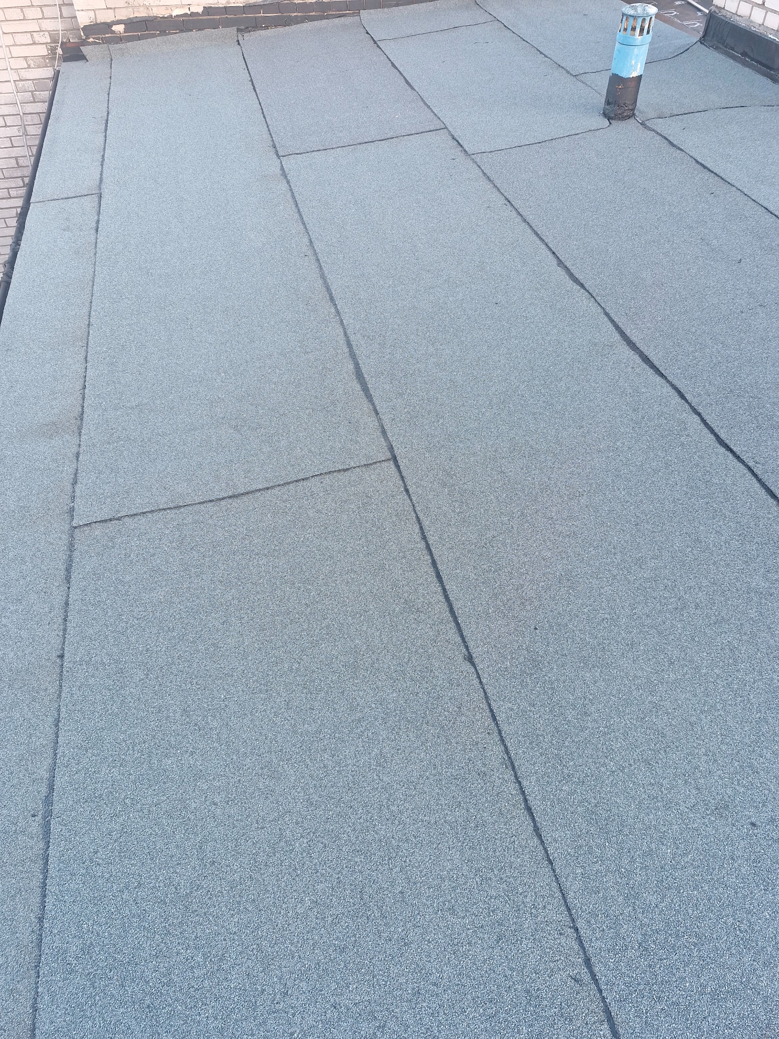 Szary, płaski dach pokryty papą termozgrzewalną z kominkiem wentylacyjnym. Widoczne łączenia papy. Ujęcie z góry, lekki kąt.