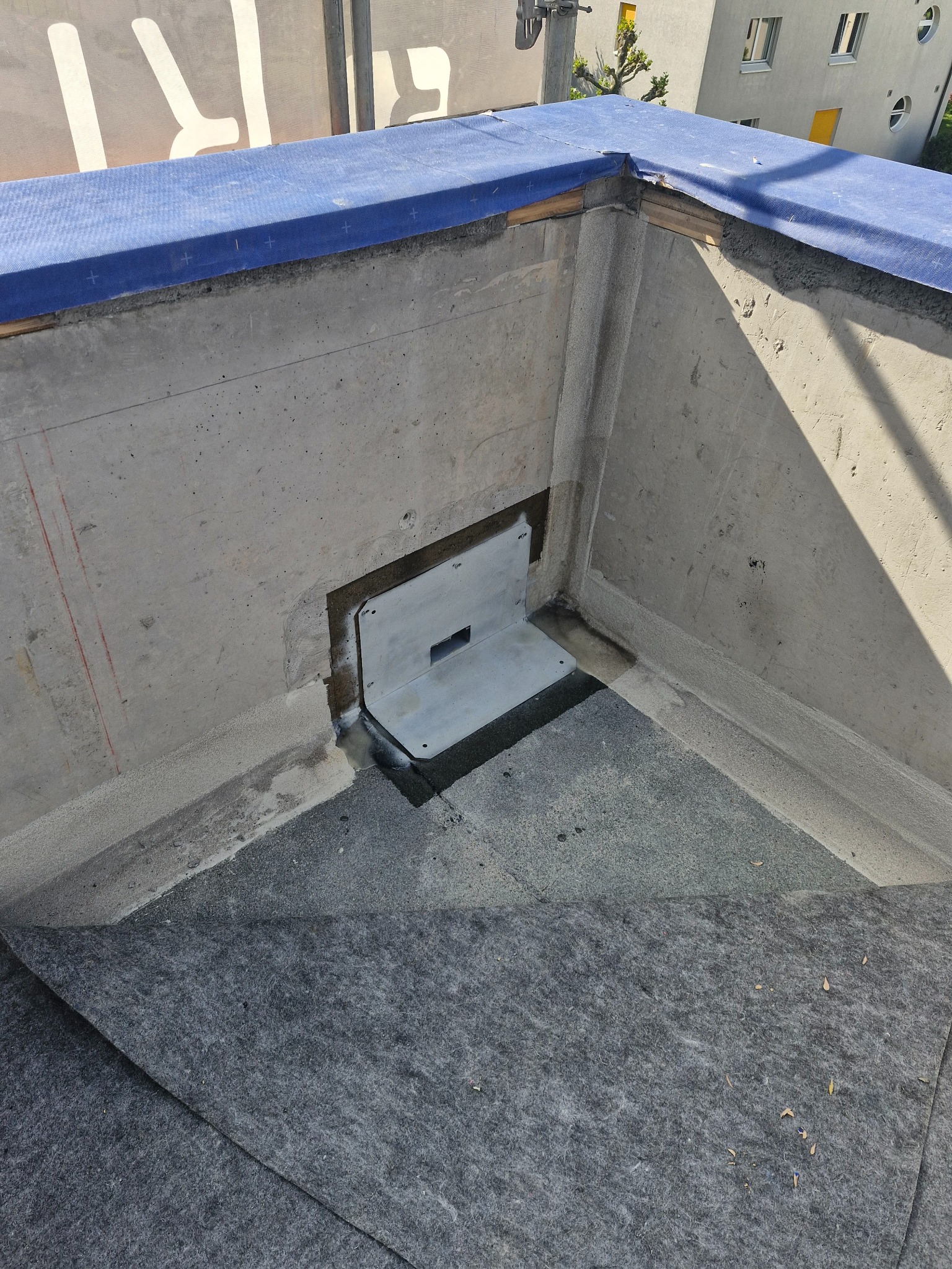 Fragment dachu płaskiego z zamontowanym odpływem wody, zabezpieczony papą. Widoczne betonowe ściany attyki i niebieska membrana. Ujęcie z góry.