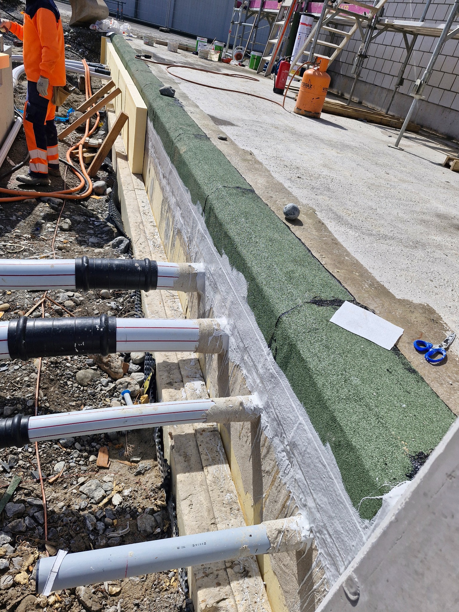 Prace izolacyjne fundamentów z widocznymi rurami, zabezpieczone białą masą uszczelniającą i zieloną membraną. W tle pracownik w pomarańczowym kombinezonie i drabina.