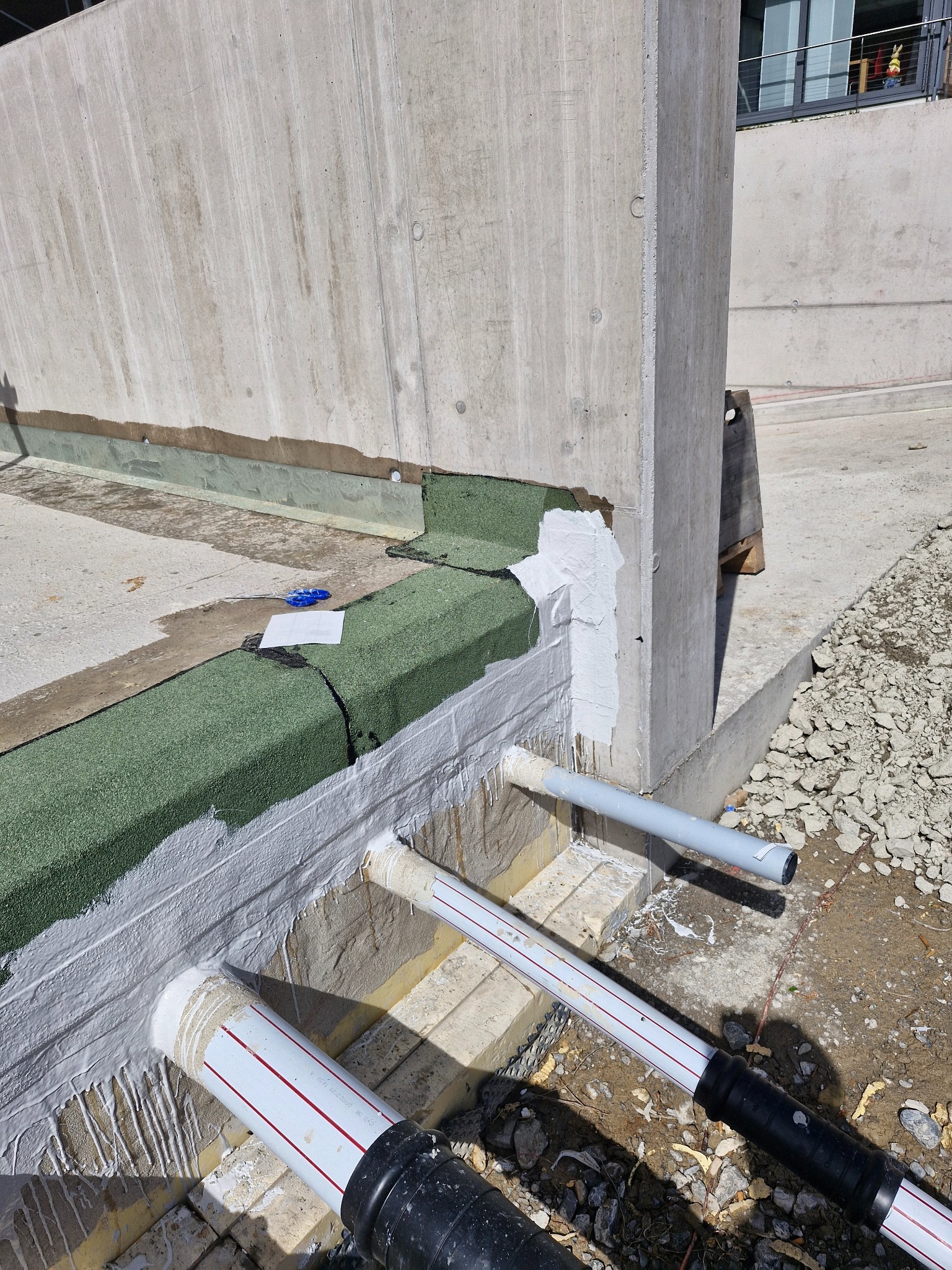 Hydroizolacja fundamentów z rurami odprowadzającymi wodę, zabezpieczona białą folią. Widoczne zielone elementy izolacyjne i betonowa ściana. Prace budowlane w toku.