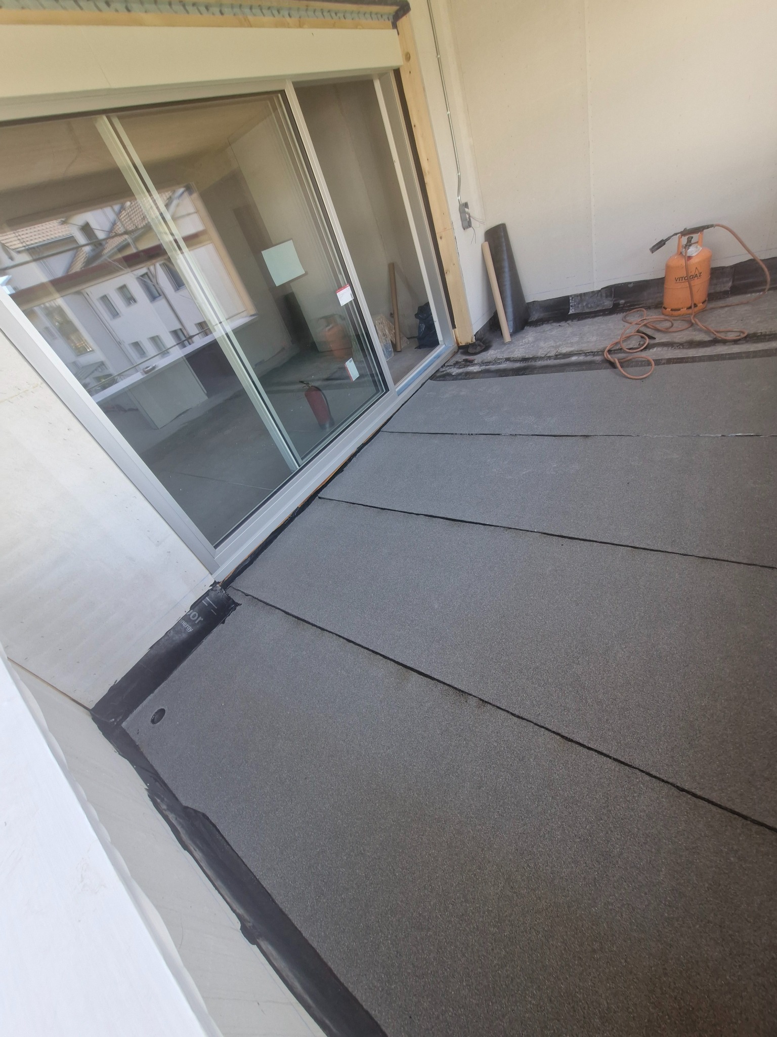 Szare płyty izolacyjne na balkonie, widoczny palnik gazowy i butla. Izolacja przeciwwodna balkonu z detalami uszczelnienia przy ścianie i oknie.