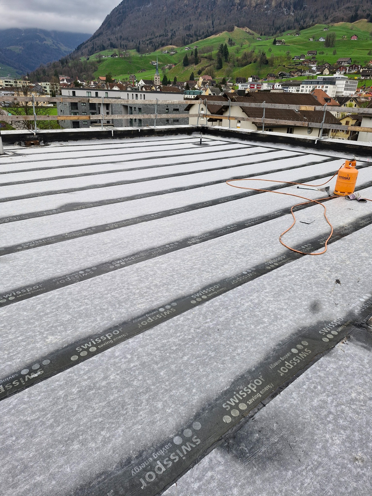 Płaski dach pokryty izolacją Swissopor, z butlą gazową i narzędziami dekarskimi. W tle zabudowa miejska i zielone wzgórza.