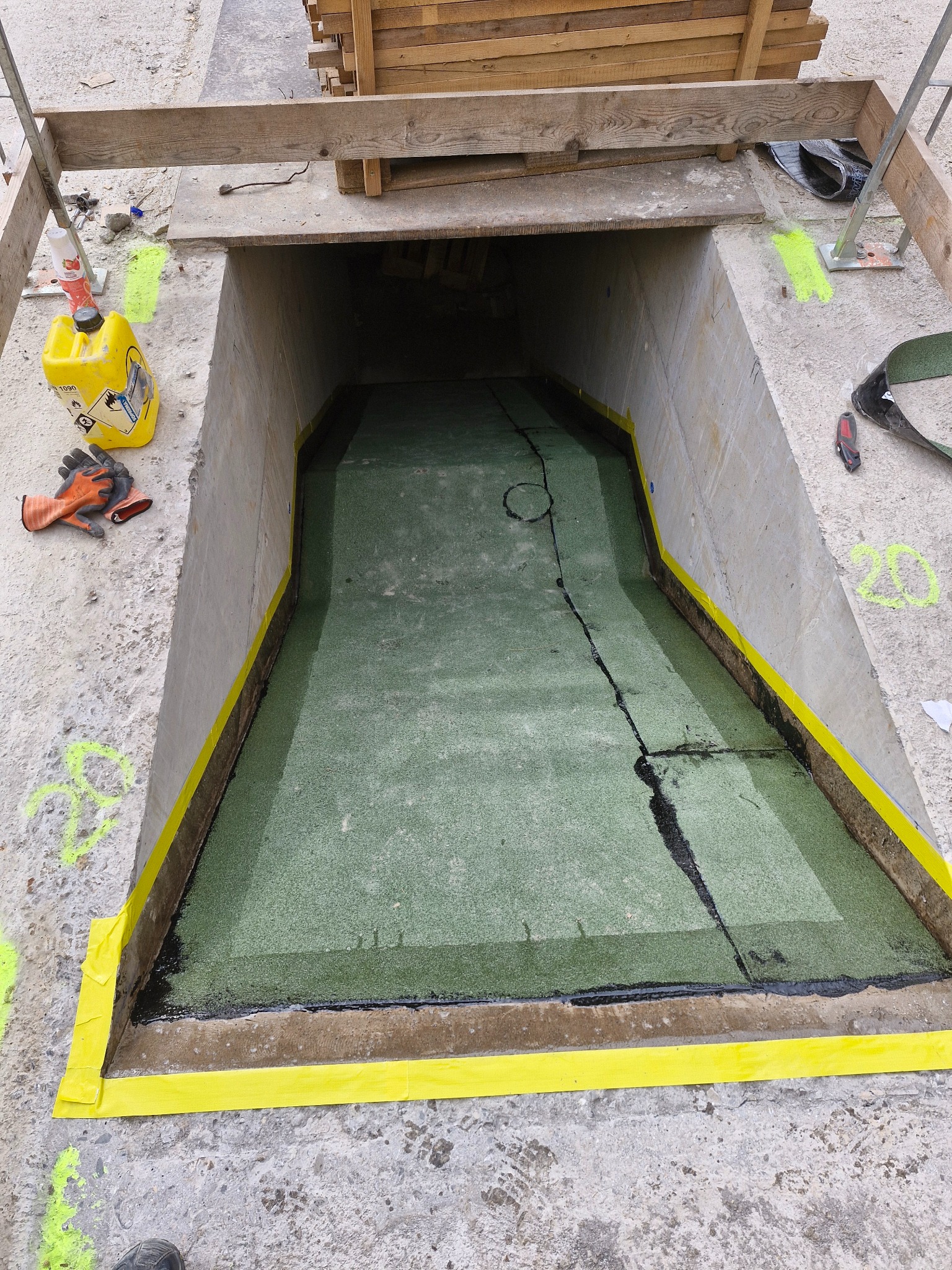 Betonowa konstrukcja z hydroizolacją wewnątrz, zabezpieczona żółtą taśmą. Widoczne narzędzia i materiały budowlane. Ujęcie z góry, podkreślające detale wykonania.