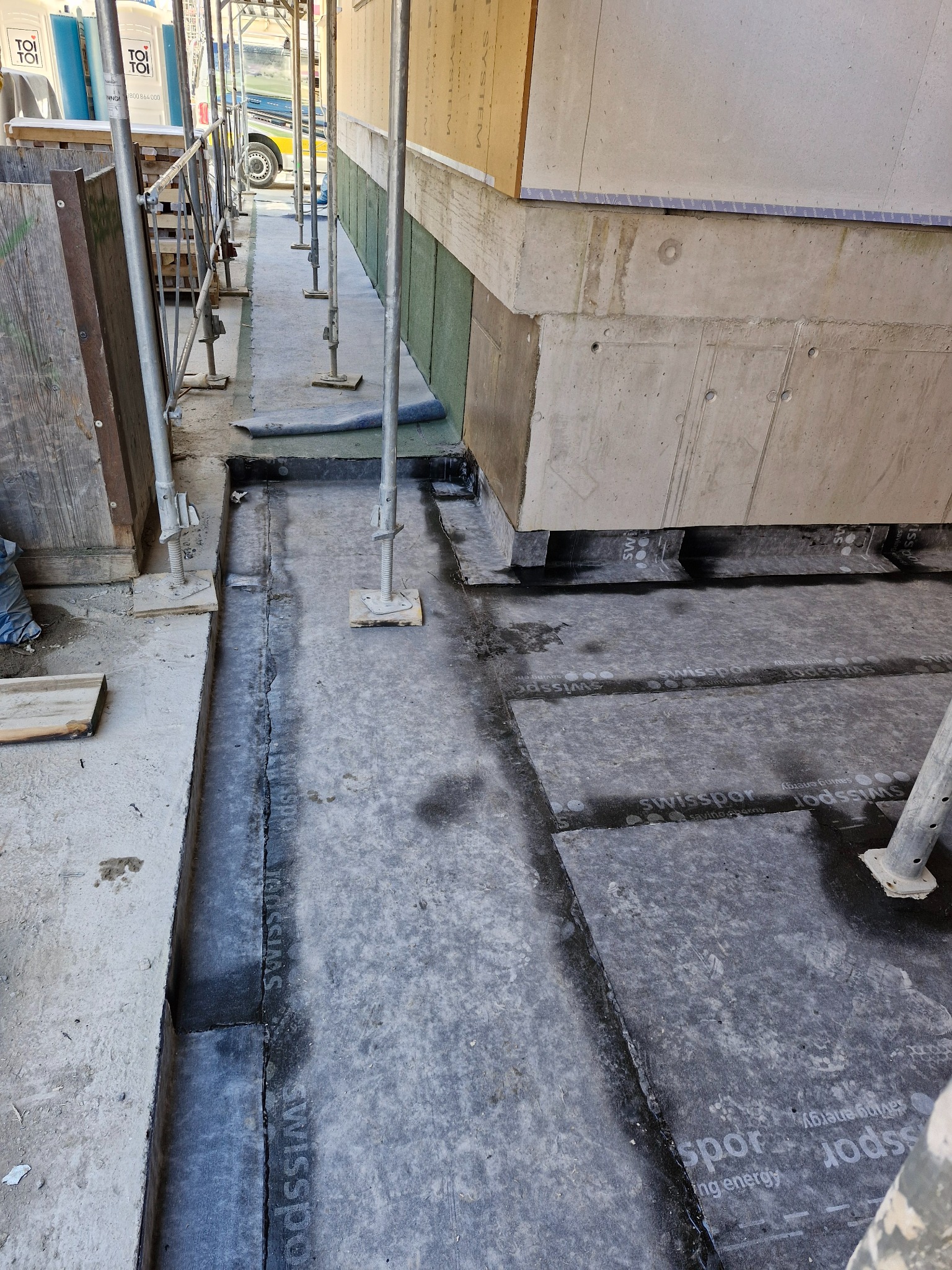 Hydroizolacja fundamentów budynku w trakcie prac. Widoczne fragmenty papy izolacyjnej z logo Swissopor, elementy rusztowania i betonowe ściany fundamentowe.