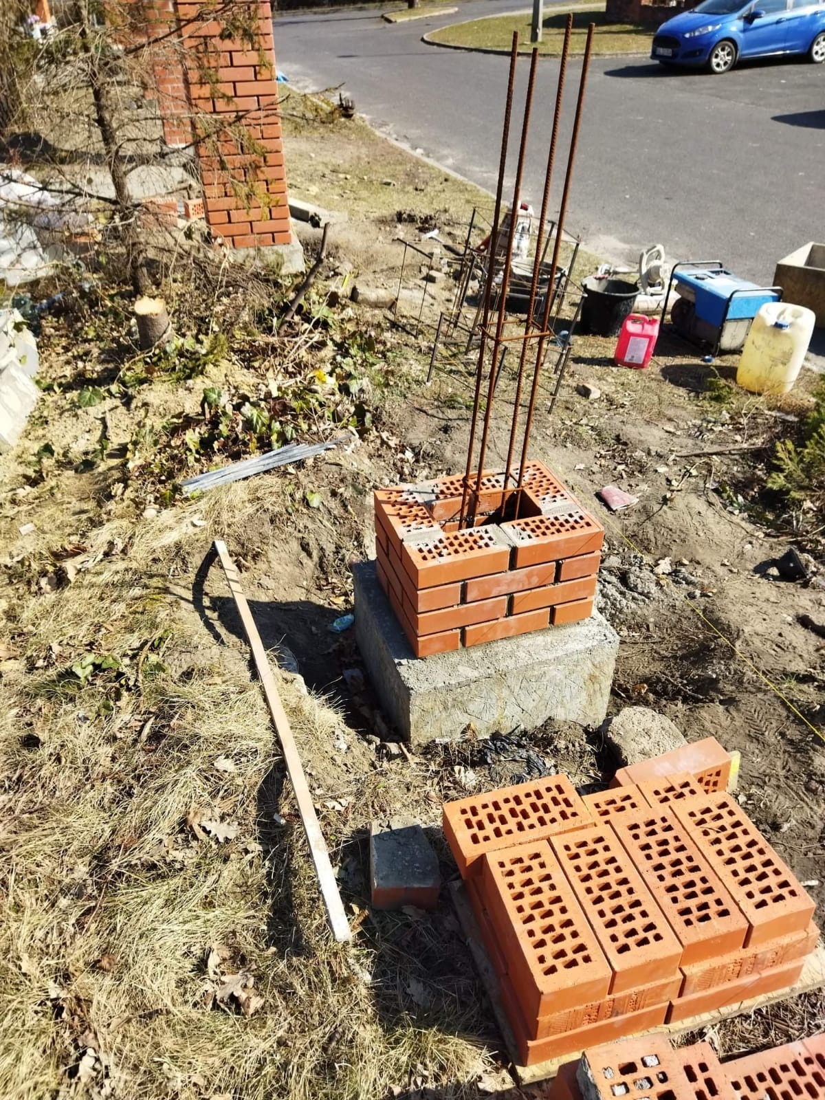 Wznoszenie fundamentu z cegły klinkierowej z wystającymi prętami zbrojeniowymi, na betonowej podstawie. Wokół gruz, cegły i narzędzia murarskie.