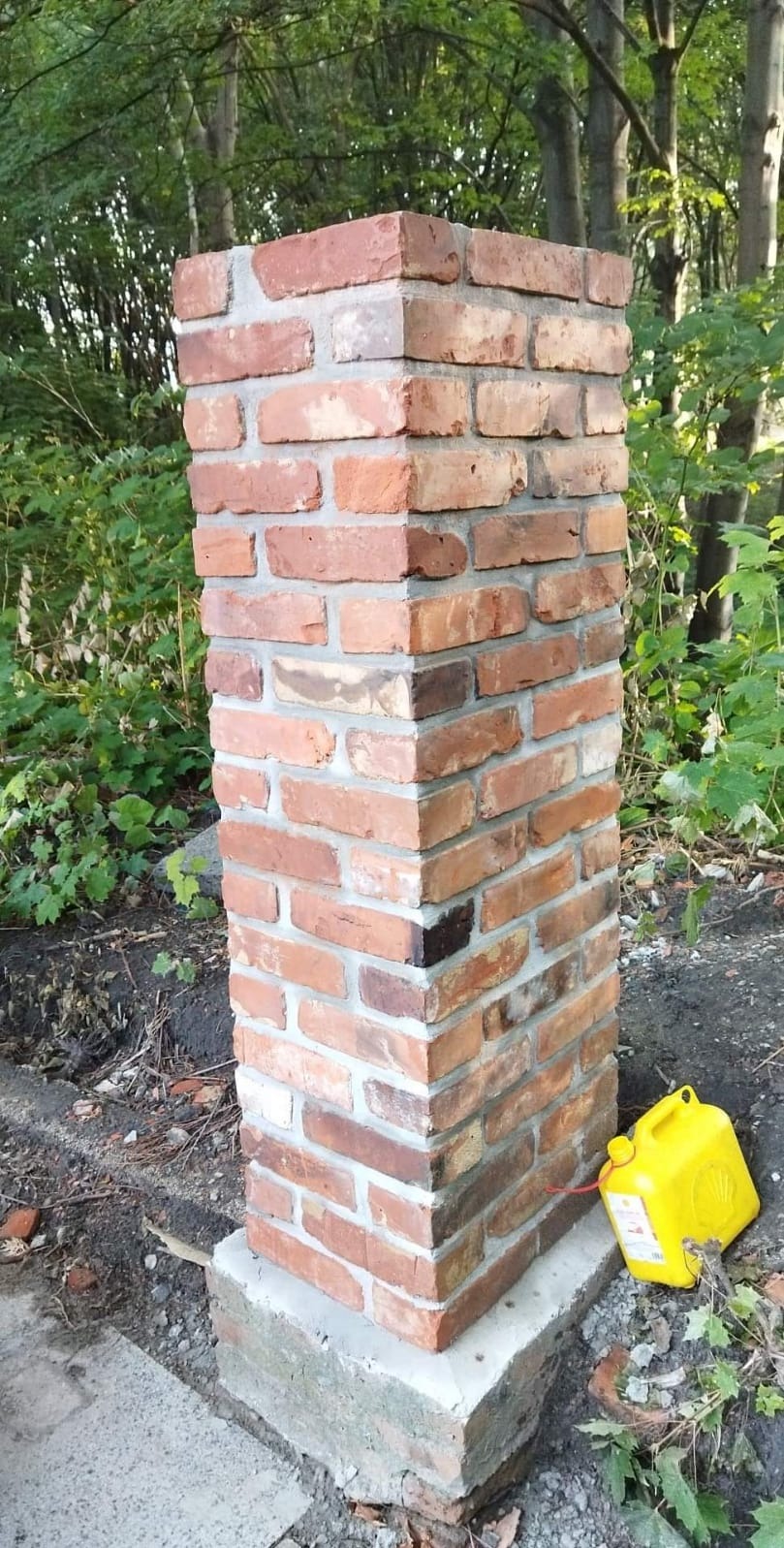 Murowany z czerwonej cegły komin wentylacyjny na betonowej podstawie, z żółtym kanistrem obok, na tle zieleni. Widoczne nierówności w ułożeniu cegieł.