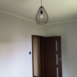 PRO - BUD - Szare ściany po malowaniu, drewniana podłoga, otwarte drzwi i lampa wisząca. Widoczny odkurzacz przemysłowy. Minimalistyczne wykończenie wnętrza po remoncie.