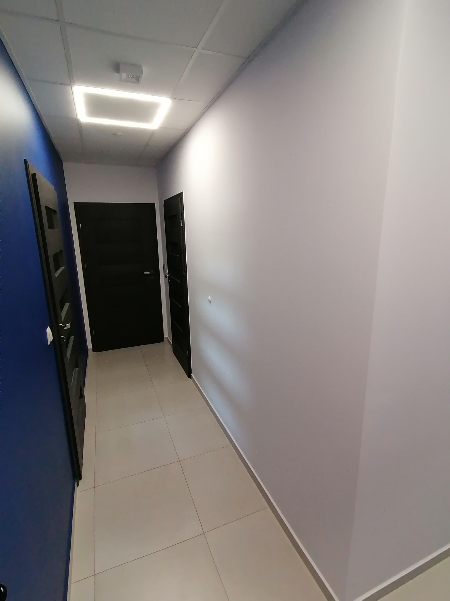 Korytarz biurowy po remoncie: świeżo pomalowane ściany w odcieniach bieli i granatu, nowe, ciemne drzwi, jasna podłoga z płytek i nowoczesne oświetlenie sufitowe.
