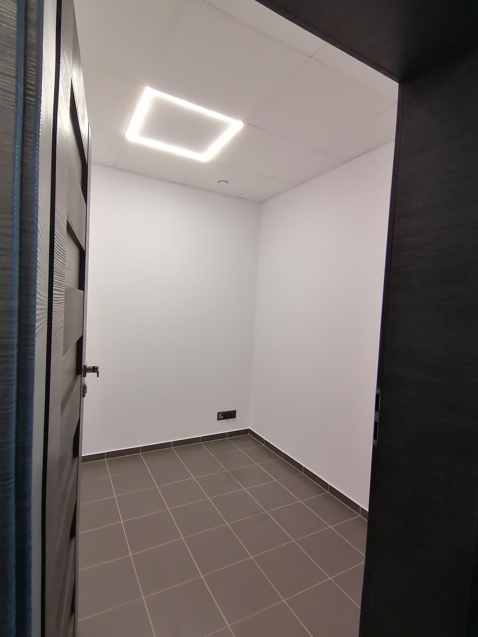 Wnętrze odnowionego pokoju z szarą podłogą w kratkę i świeżo pomalowanymi białymi ścianami. Widoczne fragmenty ciemnych drzwi po obu stronach kadru.