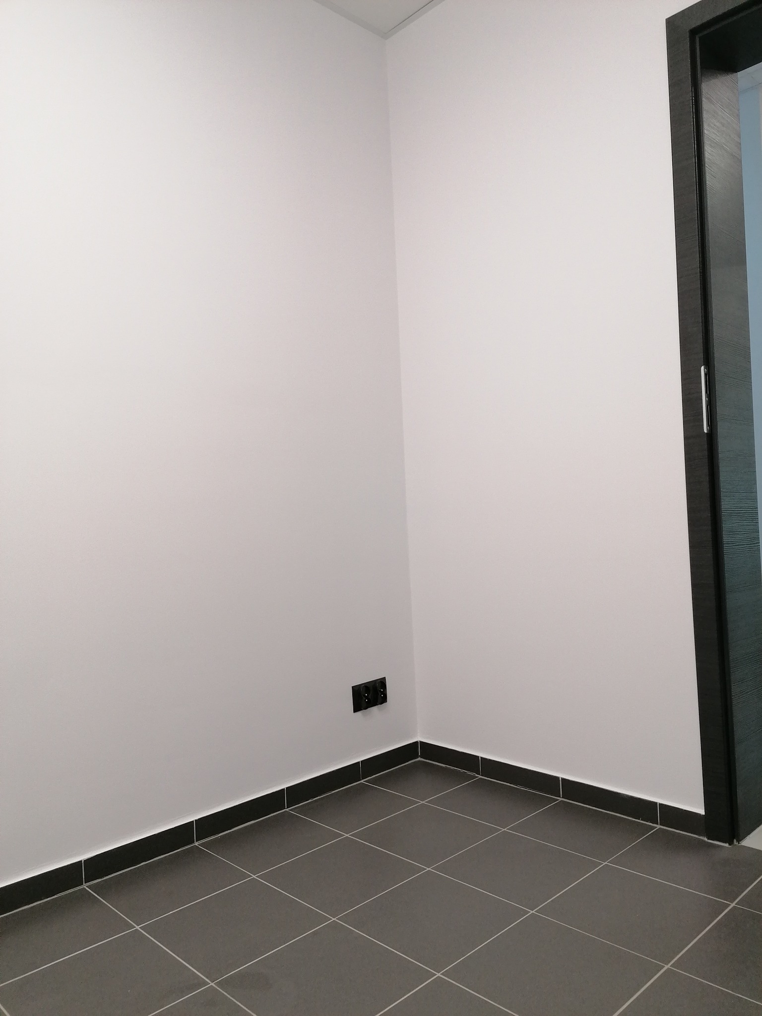 Wnętrze po remoncie: gładkie, białe ściany, szara podłoga z płytek i czarne listwy przypodłogowe. Widoczne gniazdka elektryczne i fragment czarnych drzwi.