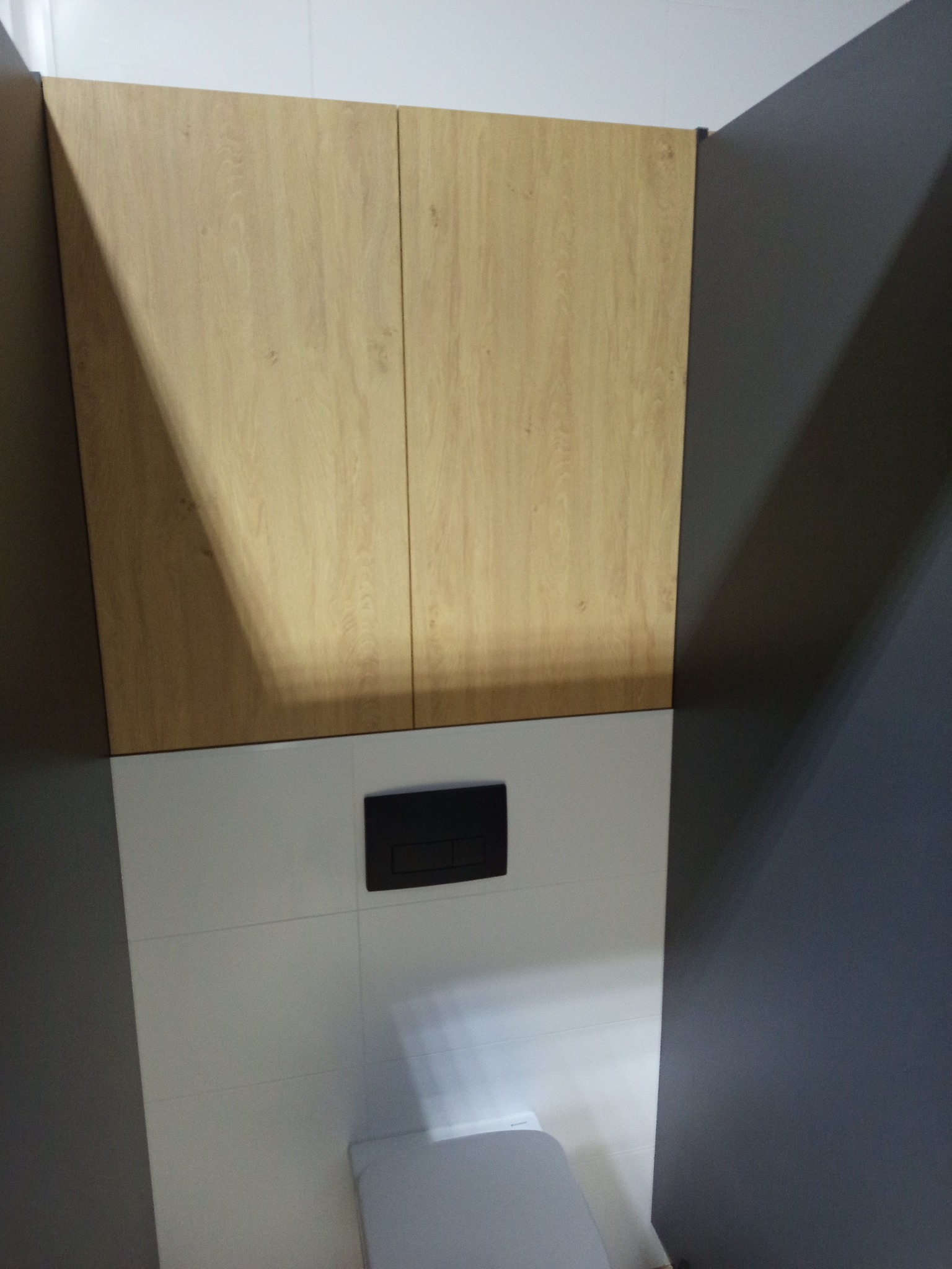 Szafka wisząca z drewnianymi frontami nad toaletą w nowoczesnej łazience z szarymi ścianami i białymi płytkami. Minimalistyczny design.