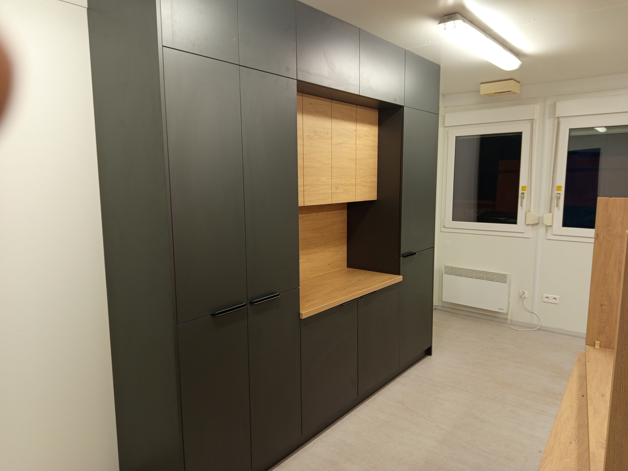 Nowoczesna szafa wnękowa z ciemnymi frontami i drewnianym blatem, w jasnym pomieszczeniu z oknami. Minimalistyczny design, proste uchwyty, widoczne oświetlenie sufitowe.