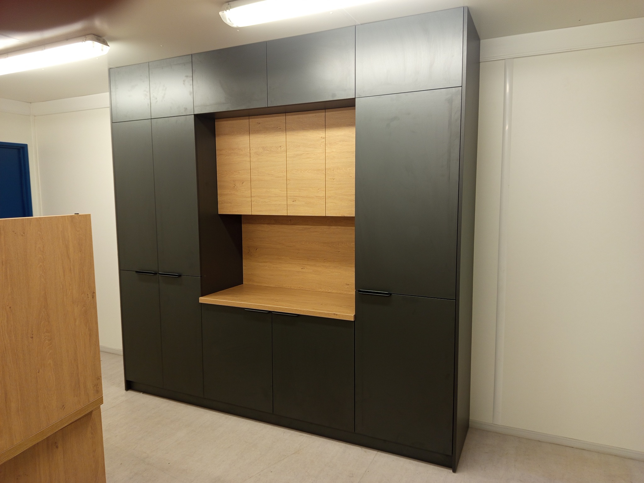 Ciemnoszara szafa wnękowa z drewnianymi elementami w ciepłym odcieniu, z minimalistycznymi uchwytami, na tle białej ściany i podłogi w jasnym odcieniu.