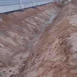 Proex Group - Wykopany rów w ziemi, zabezpieczony czarną folią, wzdłuż szarego ekranu akustycznego z zielonymi pasami. W tle widoczny wiadukt.