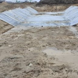 Proex Group - Wykop ziemny wyłożony folią hydroizolacyjną, zabezpieczony przez robotników w odblaskowych ubraniach. Widoczne ślady opon i błoto. Praca w toku, teren budowy.