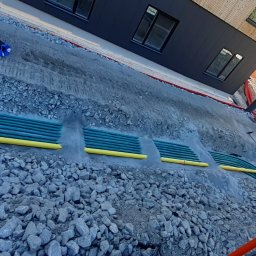 Proex Group - Wykop z ułożonymi rzędami zielonych rur z żółtymi osłonami, zasypany częściowo kamieniami, na tle budynku z ciemną i jasną elewacją. Prace ziemne.