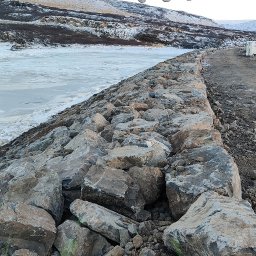 Proex Group - Brzeg rzeki umocniony dużymi kamieniami, z widocznym fragmentem łyżki koparki w górnej części kadru. W tle zaśnieżone wzgórza i częściowo zamarznięta woda.