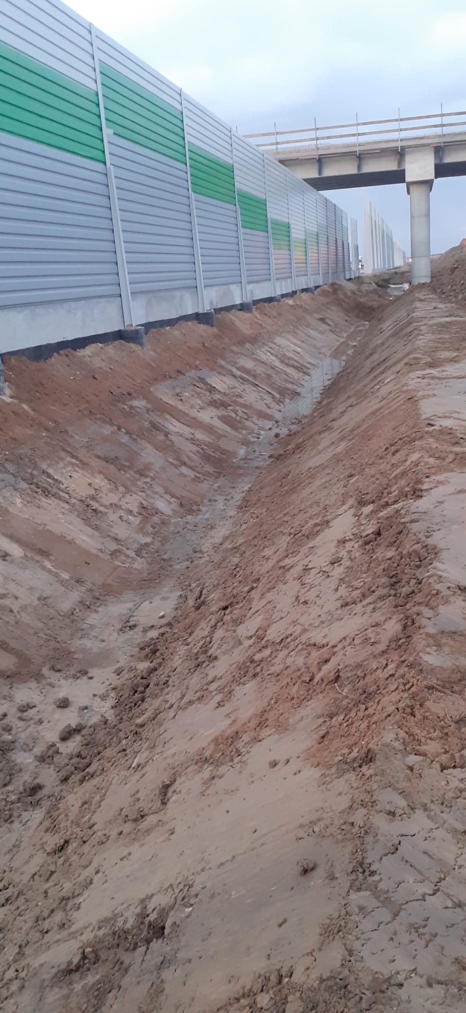 Wykopany rów w ziemi, zabezpieczony czarną folią, wzdłuż szarego ekranu akustycznego z zielonymi pasami. W tle widoczny wiadukt.