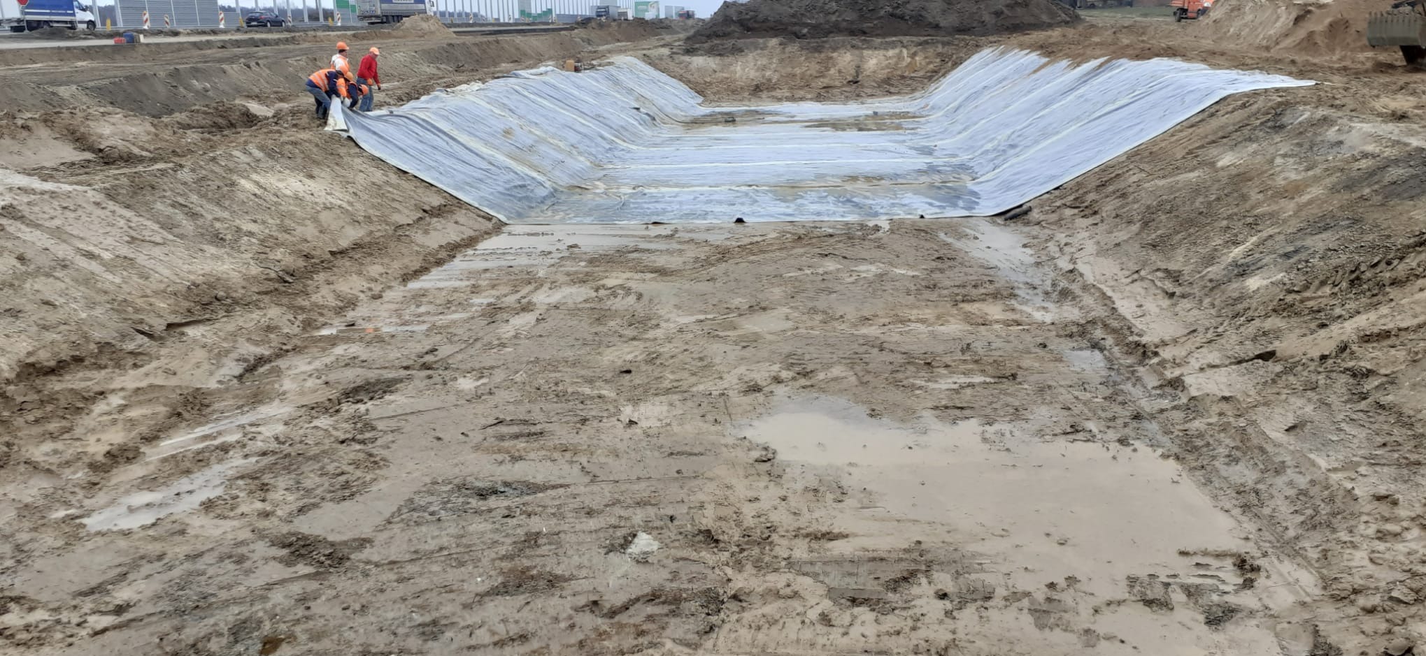 Wykop ziemny wyłożony folią hydroizolacyjną, zabezpieczony przez robotników w odblaskowych ubraniach. Widoczne ślady opon i błoto. Praca w toku, teren budowy.