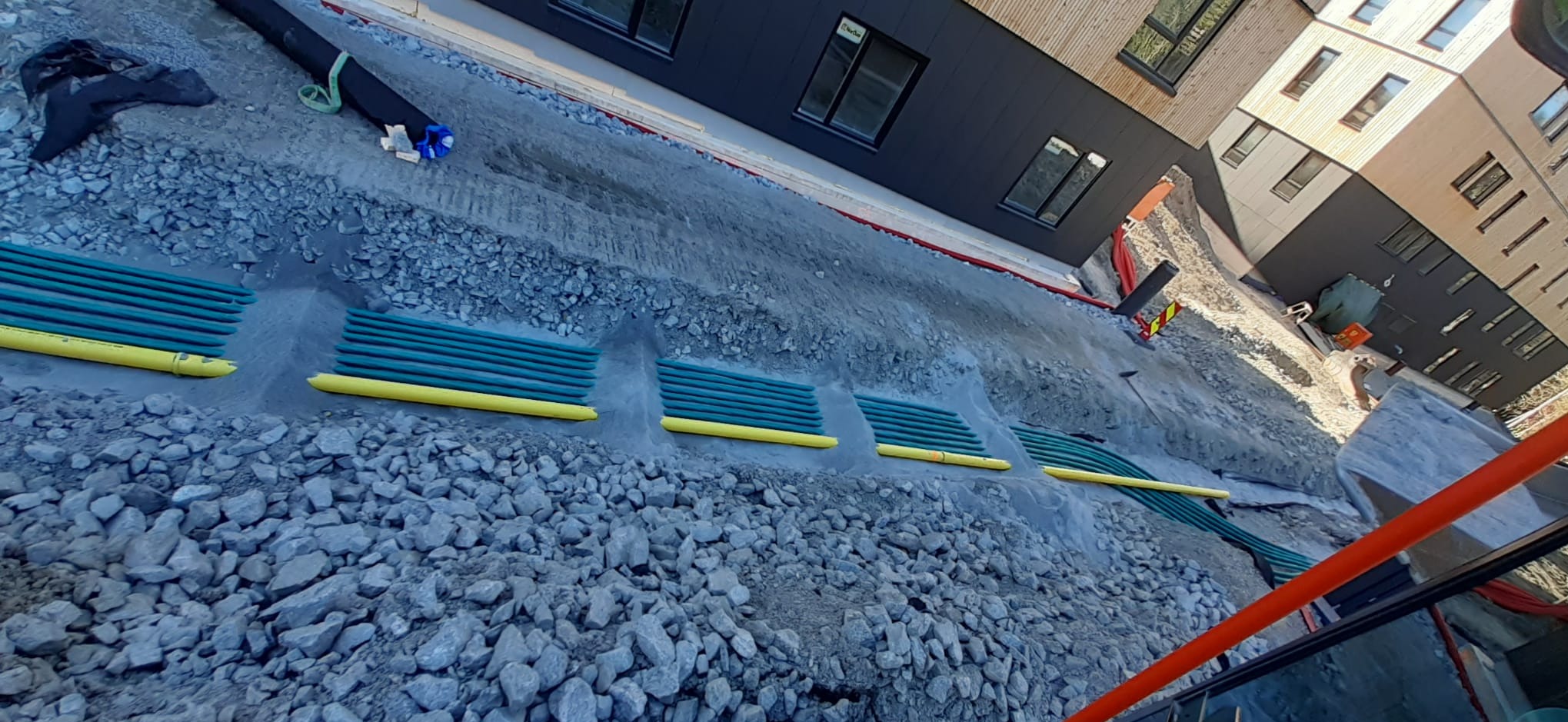 Wykop z ułożonymi rzędami zielonych rur z żółtymi osłonami, zasypany częściowo kamieniami, na tle budynku z ciemną i jasną elewacją. Prace ziemne.