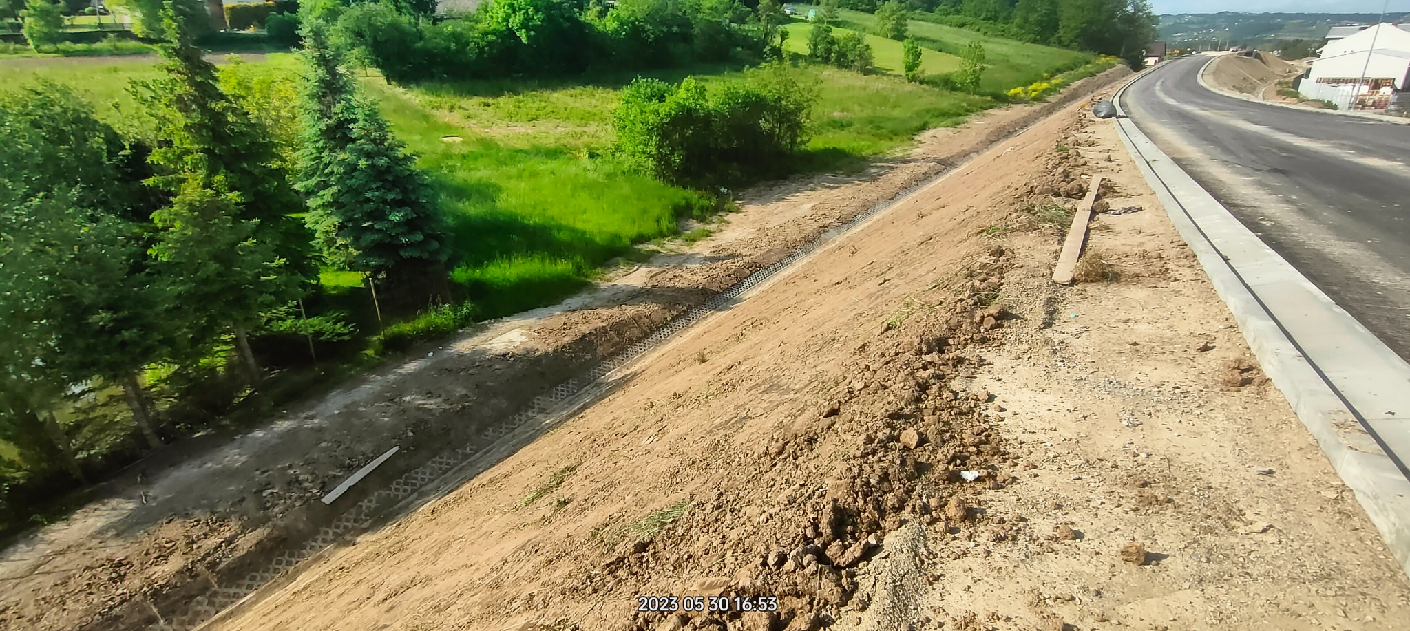 Skarpa ziemna wzmocniona geokratą, obok nowa droga z krawężnikiem. Widok z góry na teren budowy z zielenią w tle. Prace ziemne, stabilizacja gruntu.
