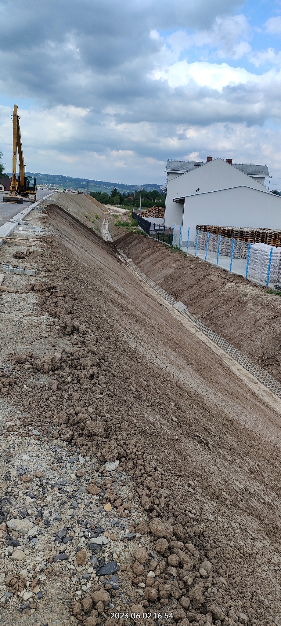 Uformowany nasyp ziemny z rowem odwadniającym, zabezpieczony ażurowymi płytami betonowymi. W tle budynek, palety i koparka. Prace ziemne w toku.