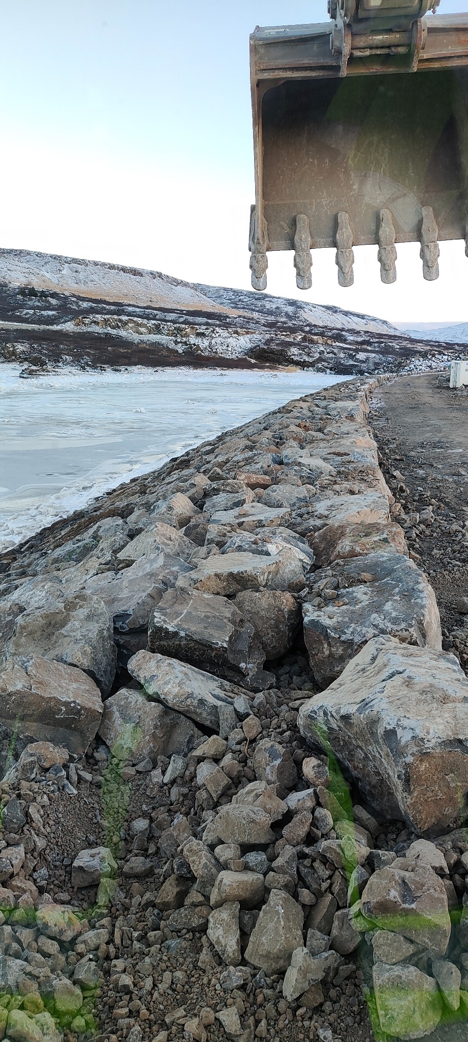 Brzeg rzeki umocniony dużymi kamieniami, z widocznym fragmentem łyżki koparki w górnej części kadru. W tle zaśnieżone wzgórza i częściowo zamarznięta woda.