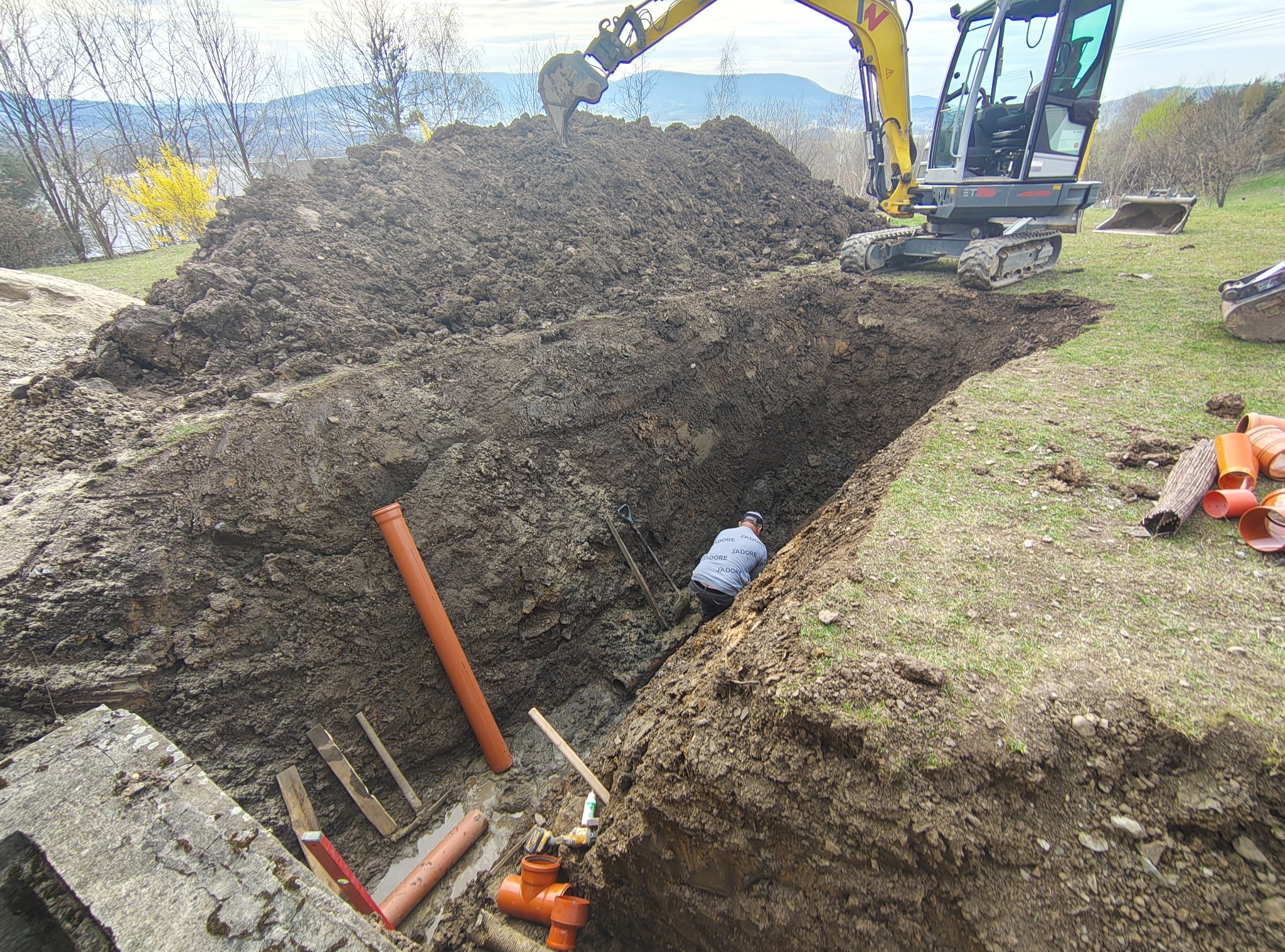 Głęboki wykop z pomarańczowymi rurami kanalizacyjnymi, pracownik w środku, koparka na górze i sterta ziemi w tle, krajobraz z drzewami i górami.