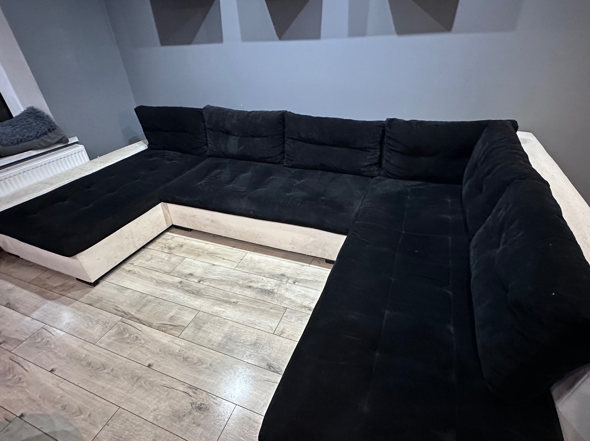 Czarna, narożna kanapa z welurową tapicerką na drewnianej podłodze. Widoczne geometryczne dekoracje na ścianie. Nowoczesny design.