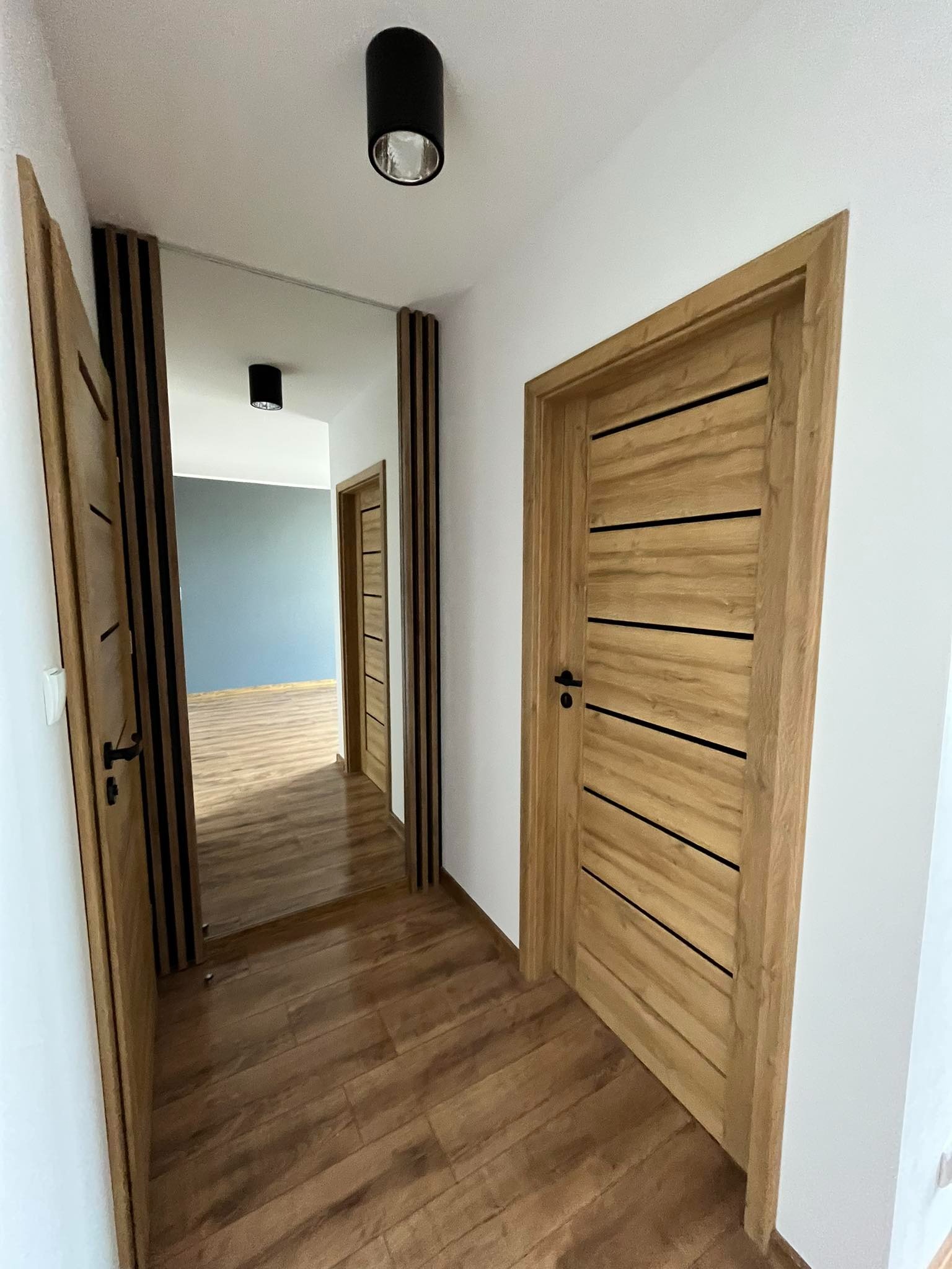 Nowoczesne, drewniane drzwi wewnętrzne z czarnymi akcentami w jasnym korytarzu z drewnianą podłogą. Widok na pomalowane na szaro pomieszczenie w głębi.