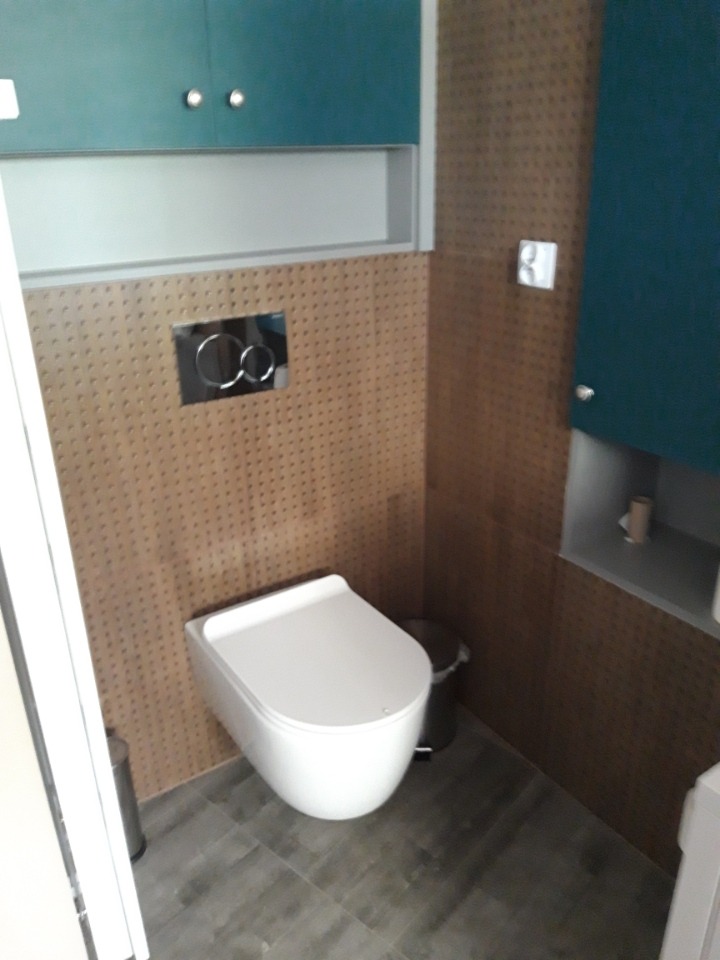 Nowoczesna toaleta podwieszana z zabudową w brązowej mozaice i turkusowymi szafkami. Minimalistyczny design, szara podłoga, chromowane detale.