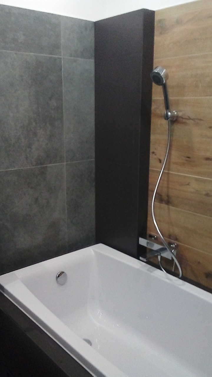 Nowoczesna łazienka z wanną, szarą płytką i drewnopodobną okładziną. Bateria prysznicowa chromowana. Minimalistyczny design, stonowana kolorystyka.