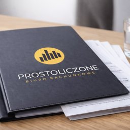 Biuro Rachunkowe Prostoliczone Małgorzata Trefler - Czarne, skórzane teczki z logo 'Prostoliczone Biuro Rachunkowe' na drewnianym biurku, obok szklanka wody i plik dokumentów. Stonowana kolorystyka, profesjonalny wygląd.