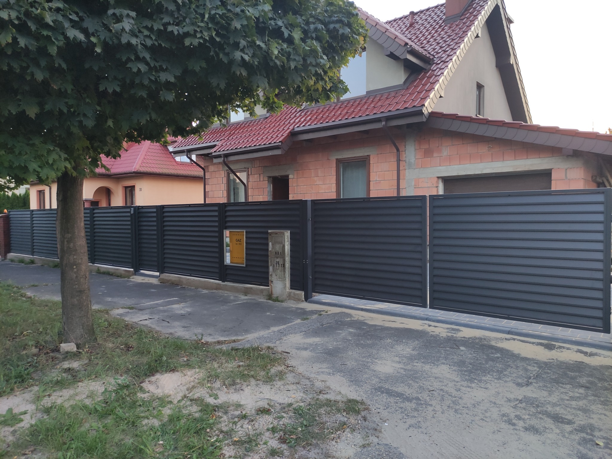 Nowoczesne, poziome, szare panele ogrodzeniowe z przesuwną bramą wjazdową, na tle domu w trakcie budowy z czerwoną dachówką i zielonym drzewem.